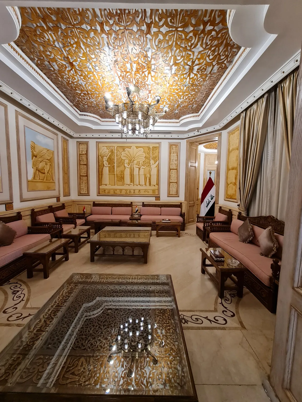 An elongated reception room with facing seating, a chandelier and a gold-detailed ceiling